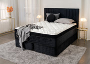 A luxury mattress on a tufted black platform bed frame, styled in a modern bedroom setting with a gray shag rug, bedside tables, and lamps.
